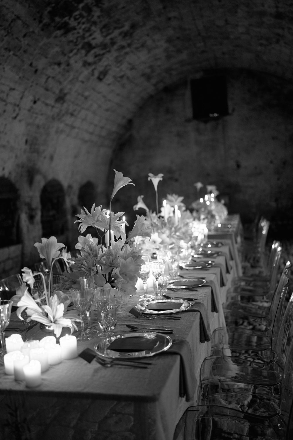 An elegant candlelit dinner table set in a rustic wine cellar with white linens and floral centerpieces.