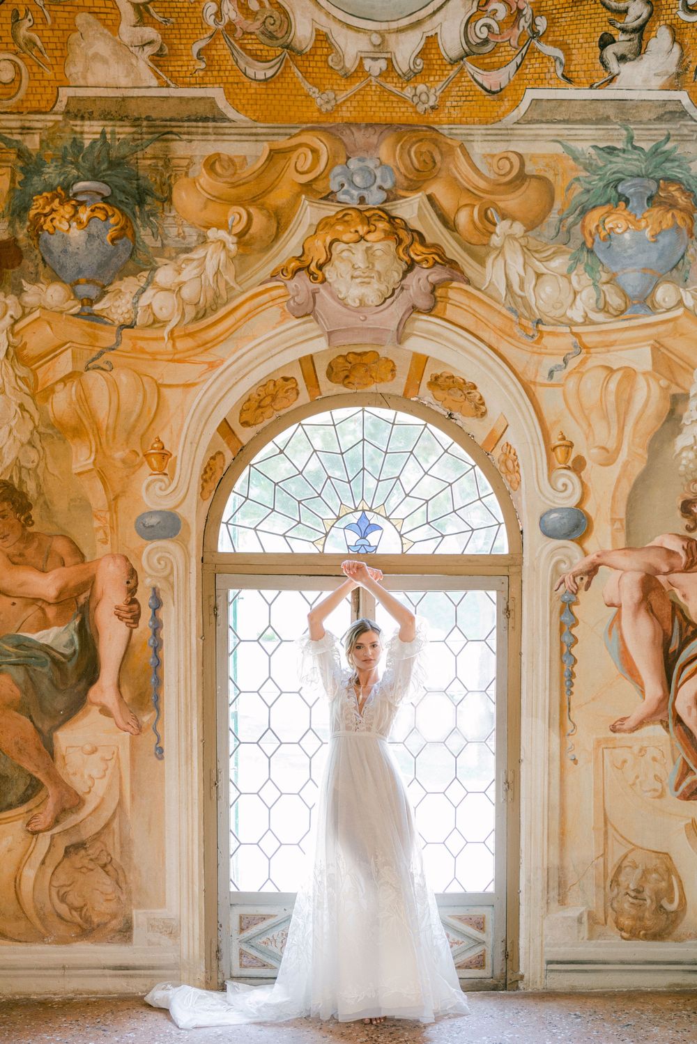 Elegant figure in white gown poses gracefully against ornate baroque window with stained glass and gilded architectural details.
