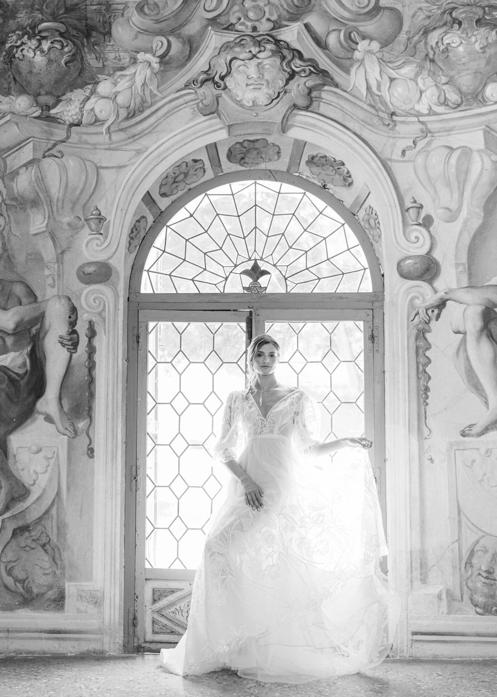 A bride in a flowing white wedding gown poses gracefully in an ornate baroque palace interior with arched windows.