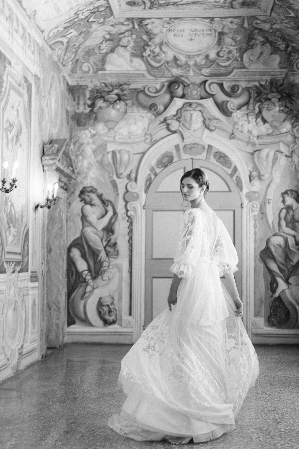 A wedding dress flows elegantly through an ornate baroque hallway with classical frescoes and decorative architecture.