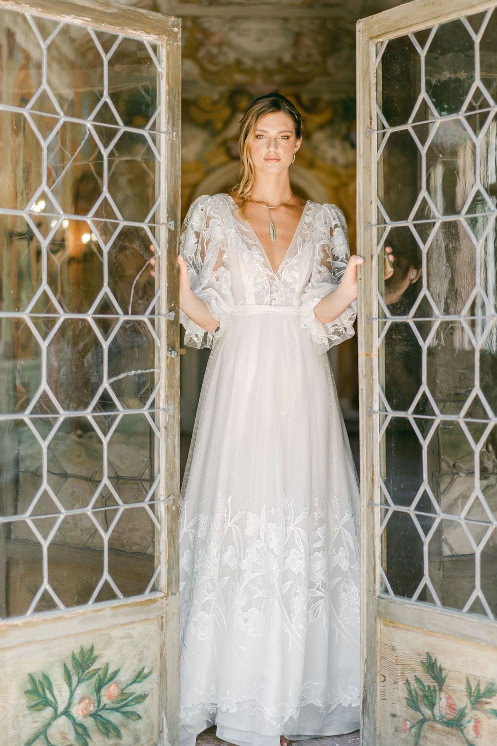 Elegant white lace wedding dress with sheer overlay and puff sleeves displayed in front of ornate lattice windows.