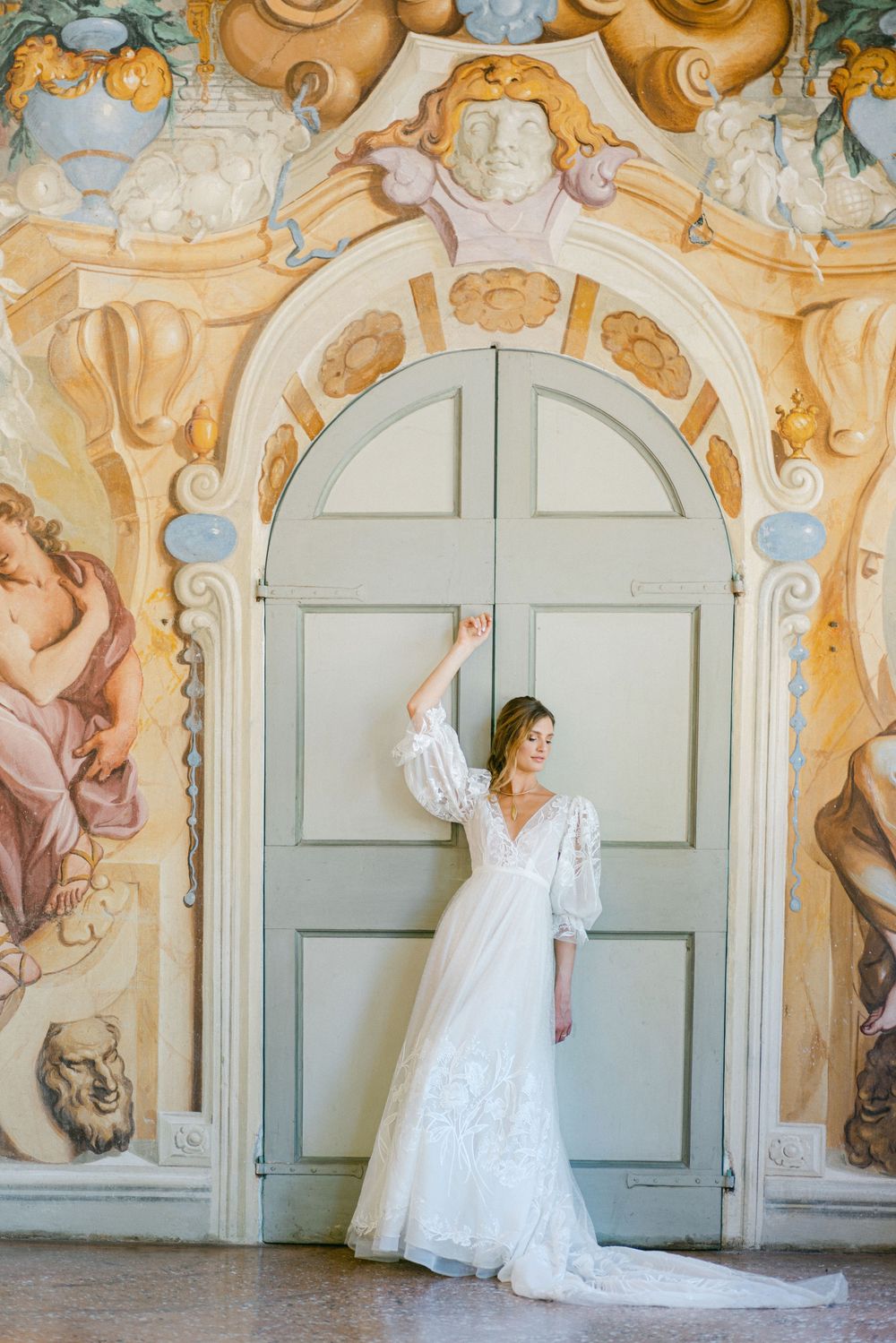 A bride in a flowing white wedding dress poses elegantly against an ornate baroque wall with painted frescoes.
