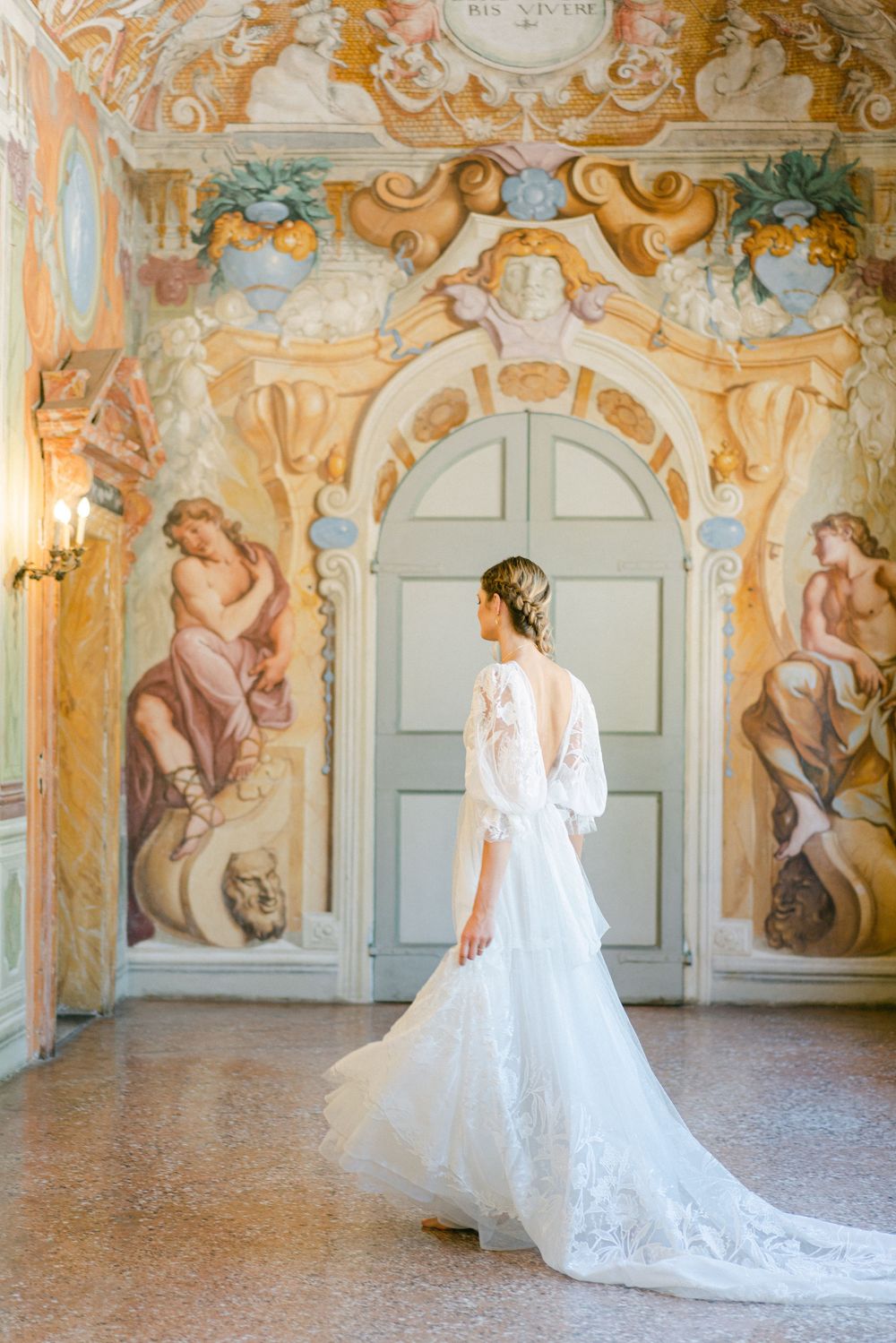 Elegant bride in white lace gown poses in ornate baroque palace room with frescoed walls and classical architecture.