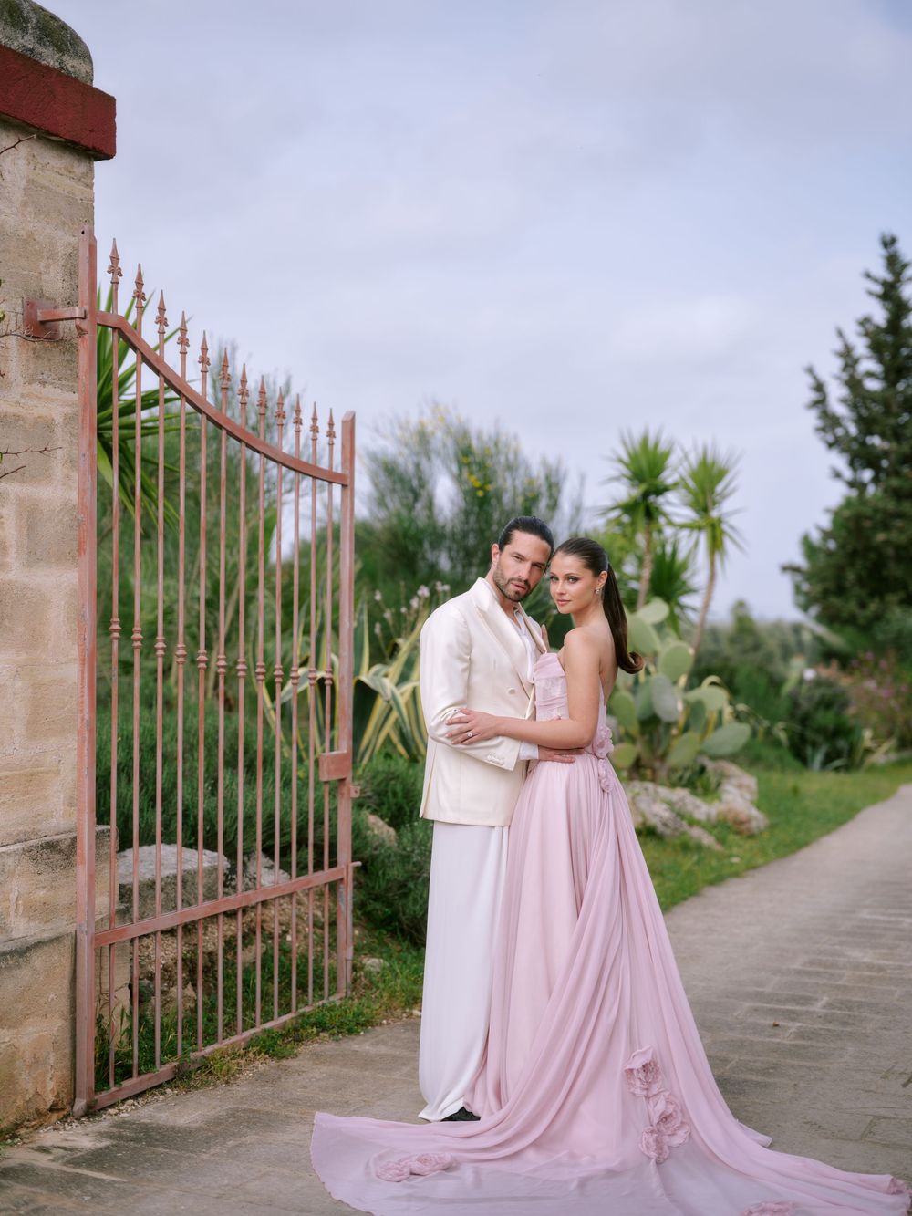A romantic couple in matching pink and white outfits pose elegantly by a rustic gate in an outdoor garden setting.