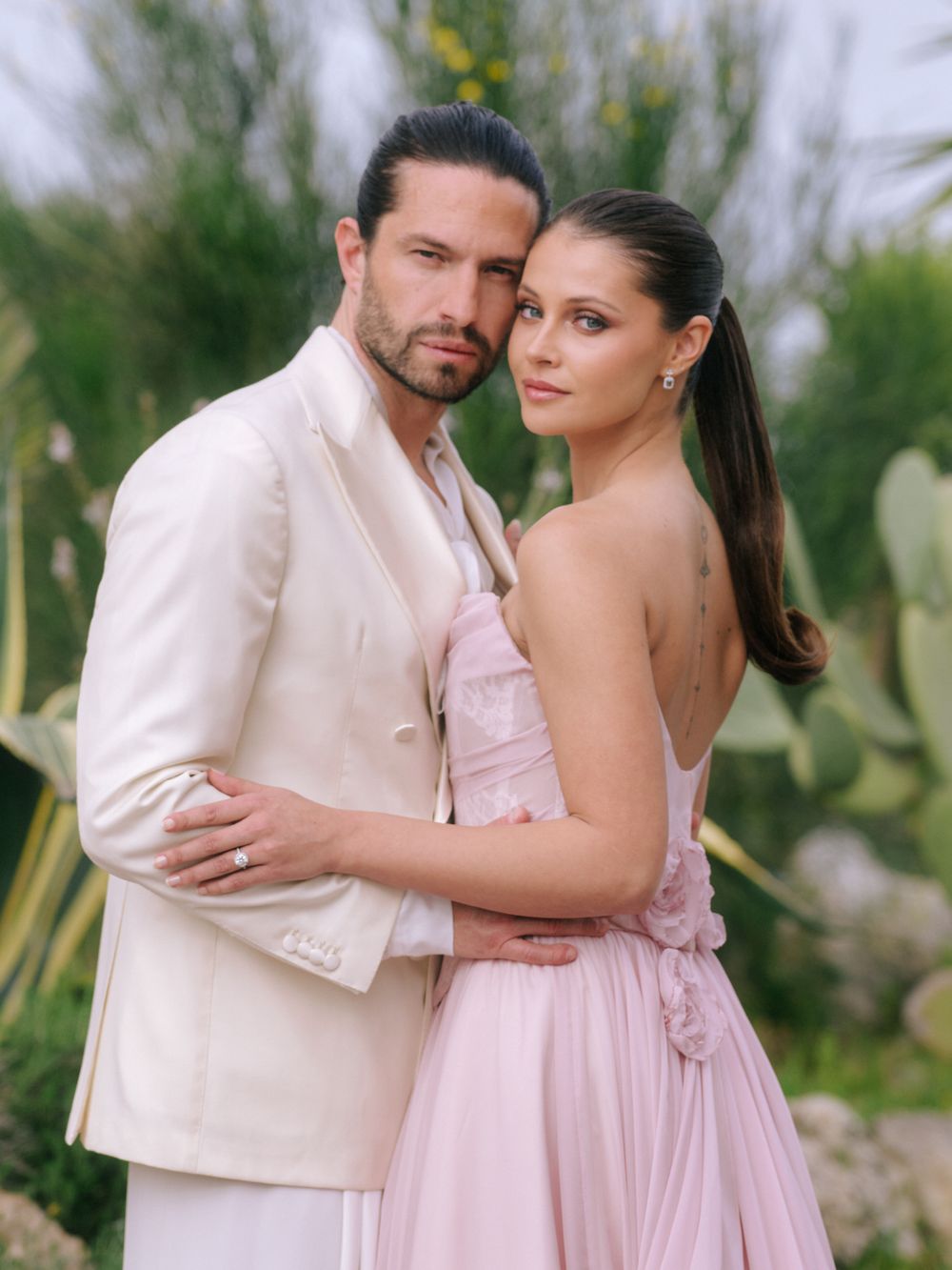 A couple in romantic poses against a desert landscape backdrop, wearing white and pink formal attire.