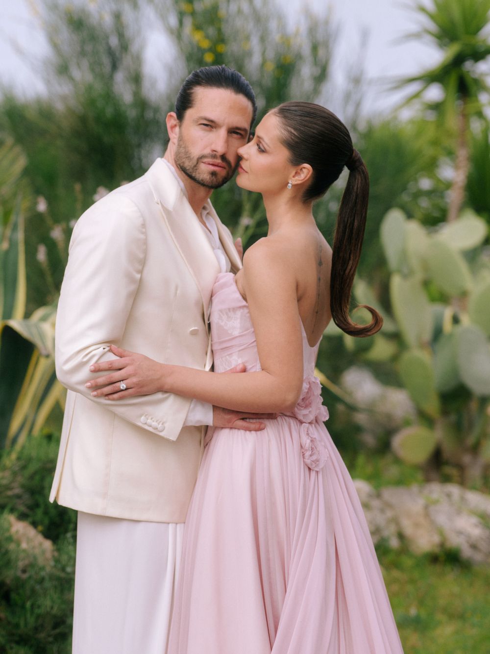 A romantic couple embraces outdoors in formal attire, with her in a pink gown and him in a beige suit amid desert plants.