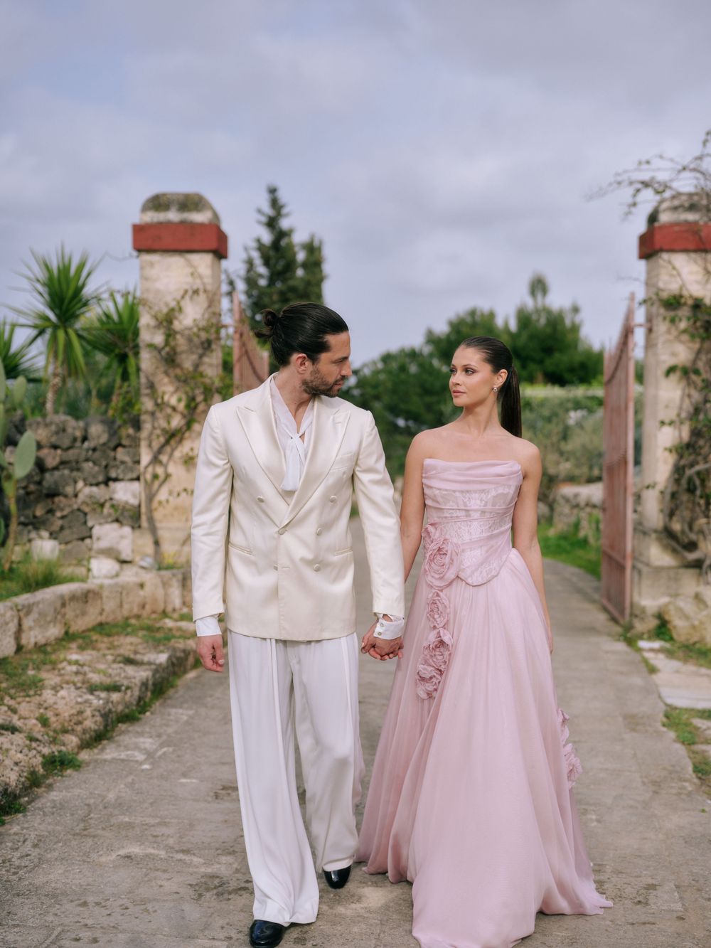 A couple in formal attire walks along a stone path with pink and white outfits in a romantic outdoor setting.