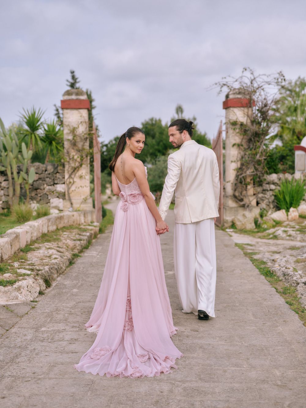 A couple in pink and white formal attire walk hand in hand along a rustic stone path during their destination wedding. Puglia wedding