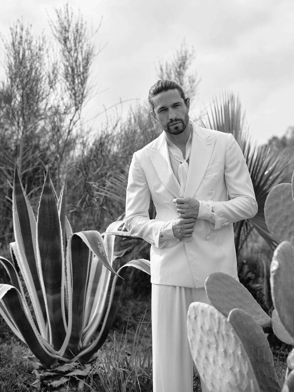 A couple in white formal wear poses among desert cacti and agave plants in a black and white photograph.