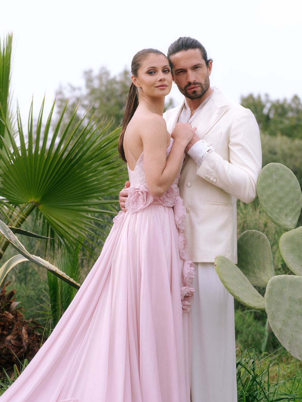 A couple poses romantically against a backdrop of palm fronds and cacti in a desert garden setting.
