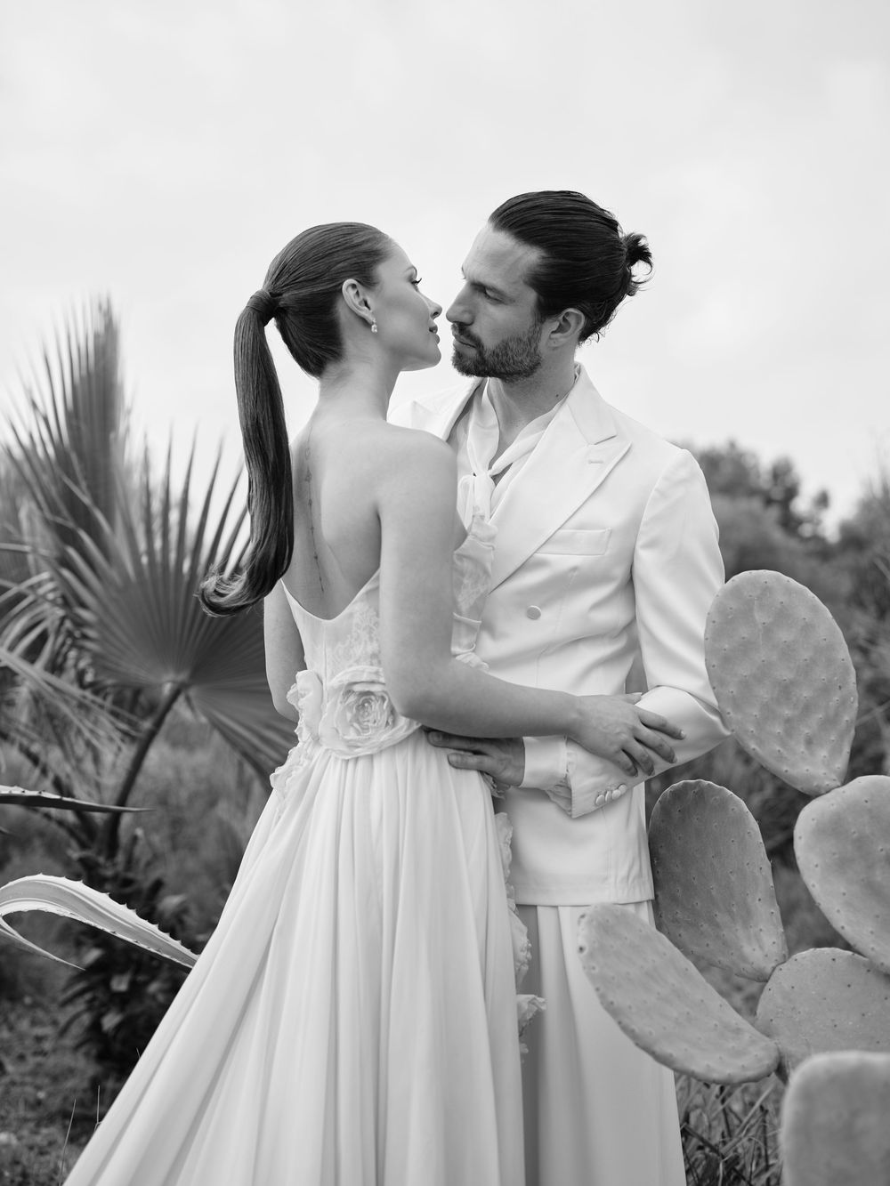 A romantic black and white photo series showing a couple in white outfits embracing in a tropical garden setting.