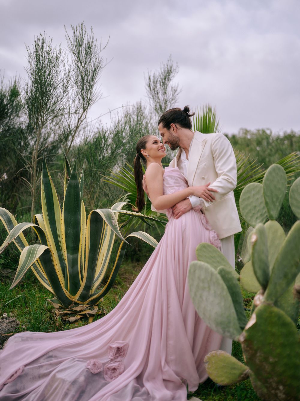 A romantic couple in formal attire embraces among desert plants and cacti during a dreamy outdoor photoshoot. Wedding in Puglia