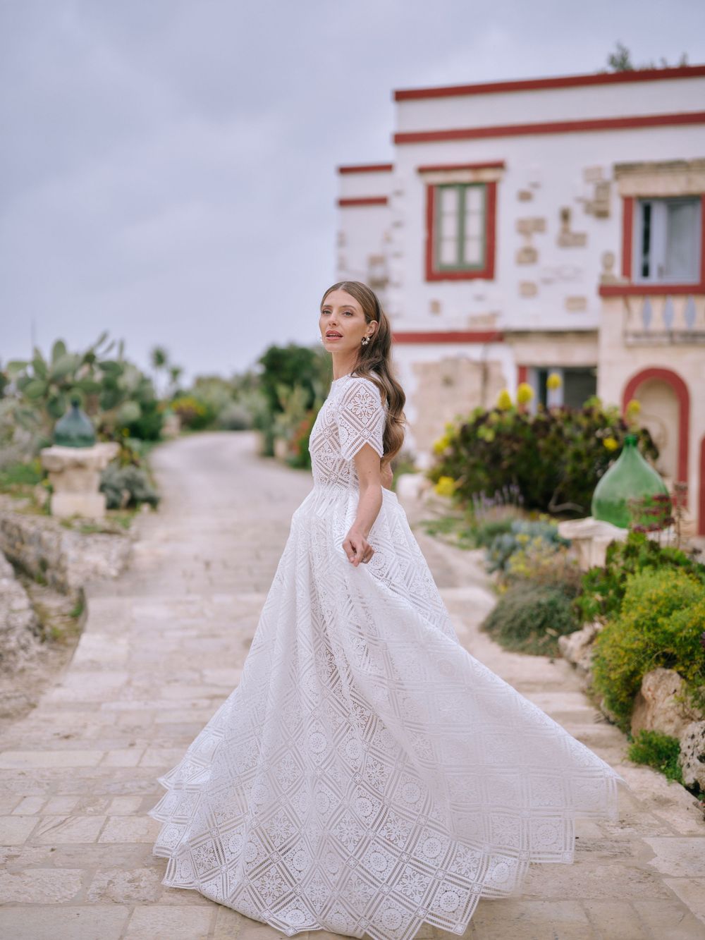 A bride in a flowing lace wedding dress twirls gracefully on a stone patio surrounded by Mediterranean-style architecture. Puglia wedding