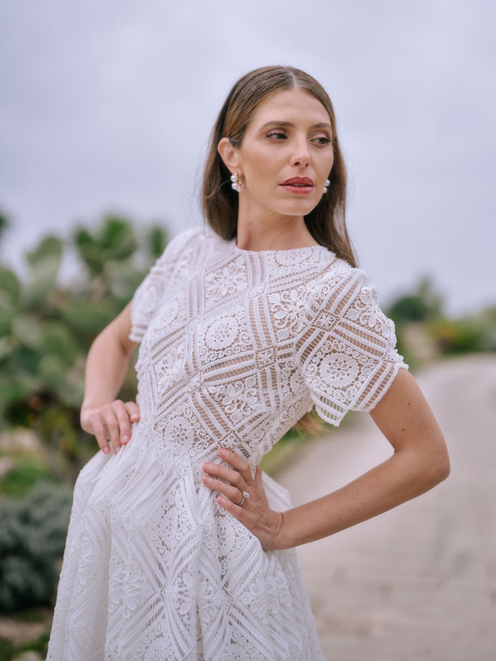 A white lace wedding dress with geometric patterns and short puff sleeves is modeled against a soft outdoor background.