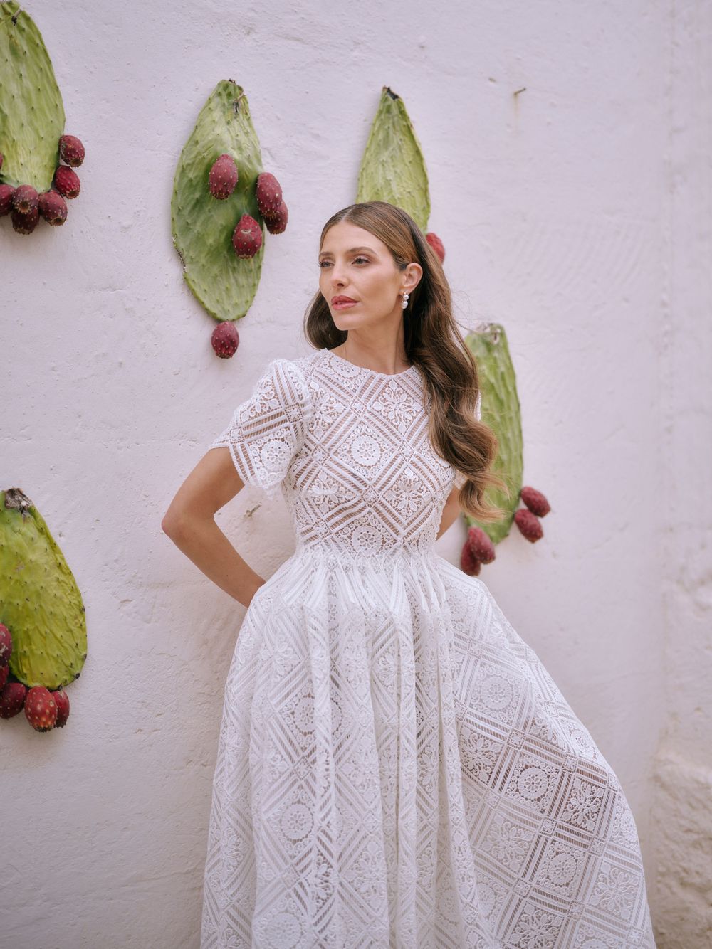 A model in a white lace dress poses against a rustic wall decorated with cactus paddles and red prickly pears. Puglia wedding photographer