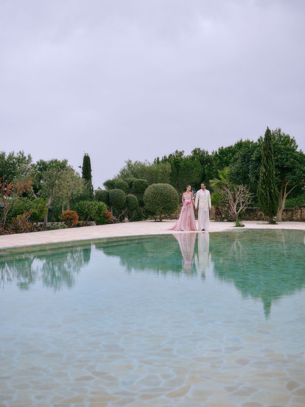 A serene poolside setting with elegant figures reflected in turquoise water surrounded by lush Mediterranean gardens. Wedding photographer