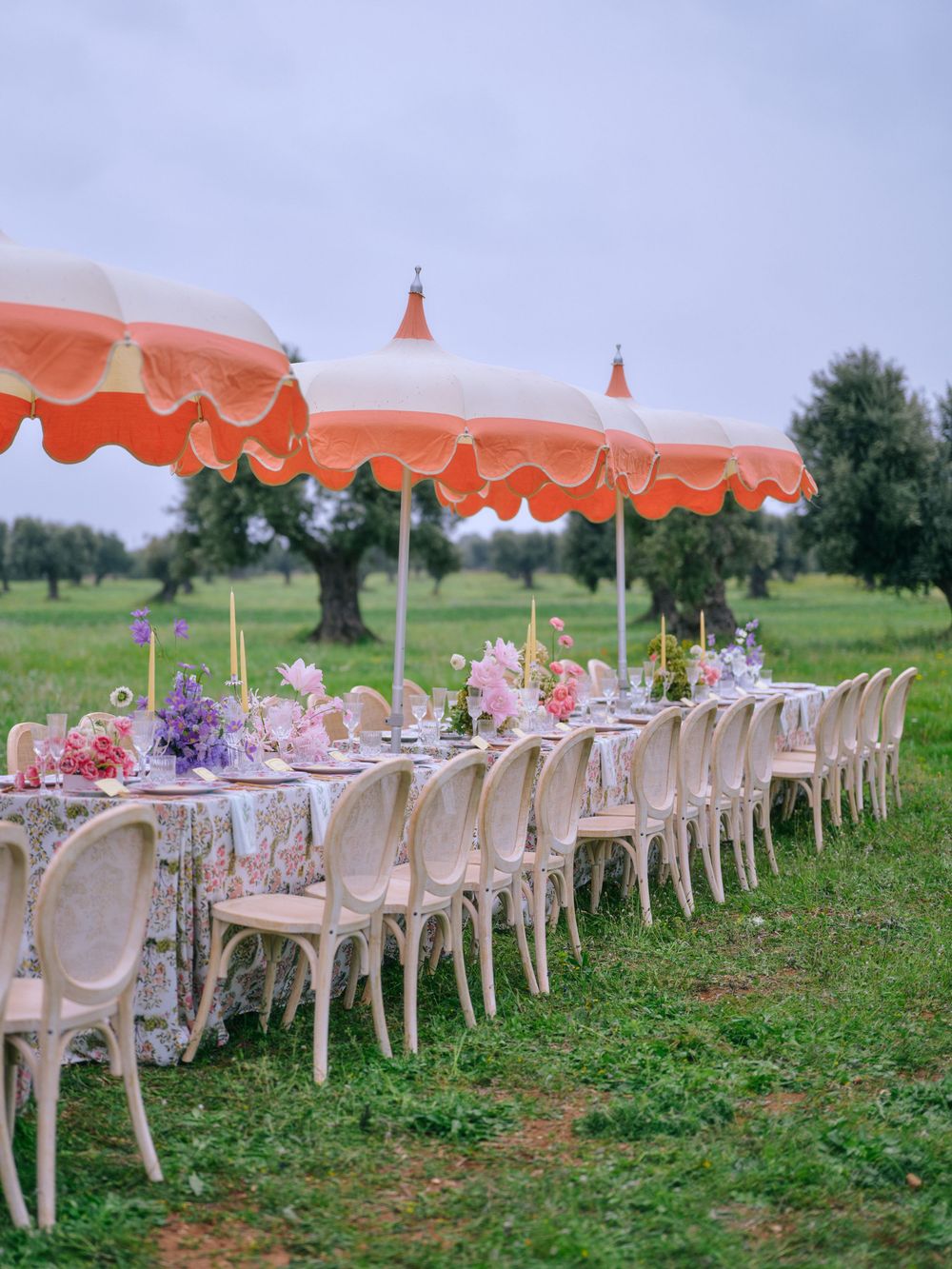 Outdoor wedding reception setup with coral umbrellas and white chairs arranged in rows on a grassy lawn.