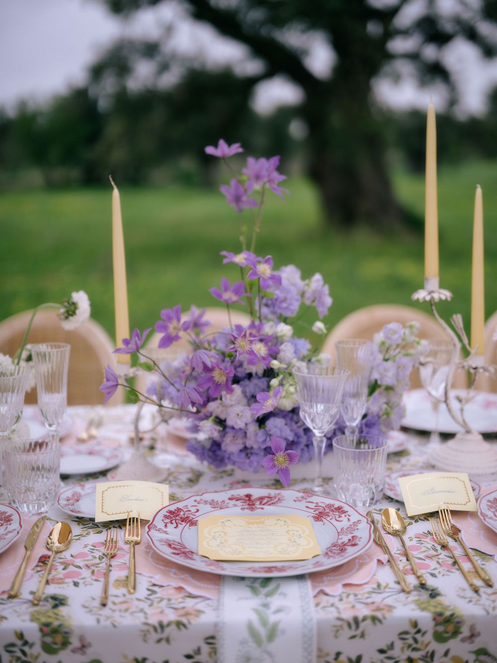 An elegant wedding table setting with purple flowers, tall candles, and vintage-style floral tablecloth in an outdoor garden.