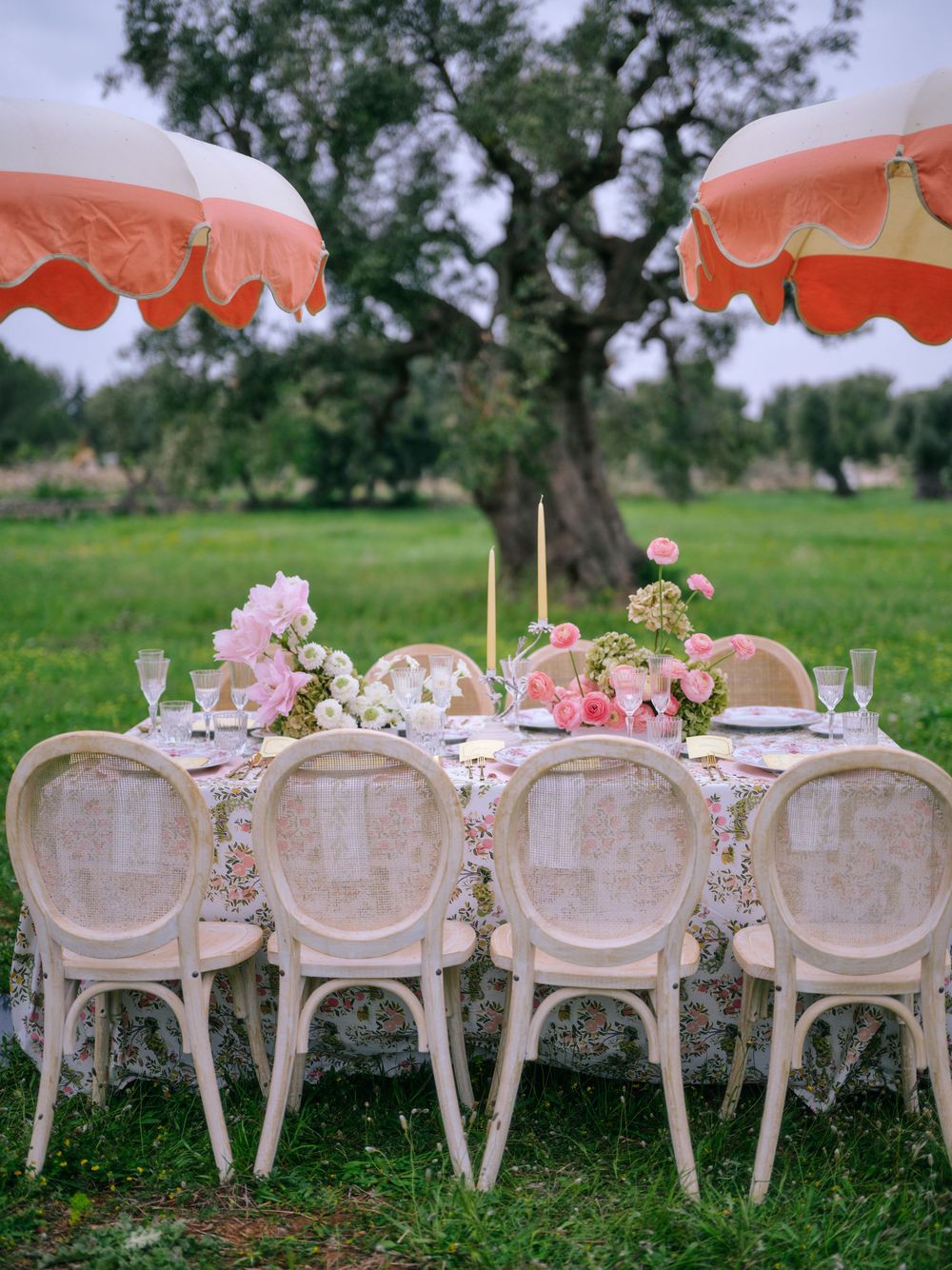 Elegant outdoor table setting with pink flowers, vintage cream chairs and coral umbrellas in a garden setting.