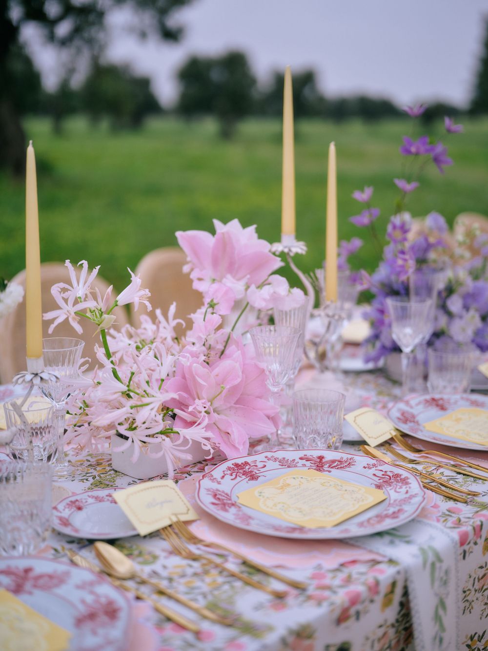 Romantic garden party table decorated with pink and purple flowers, gold candles, and vintage-style place settings.