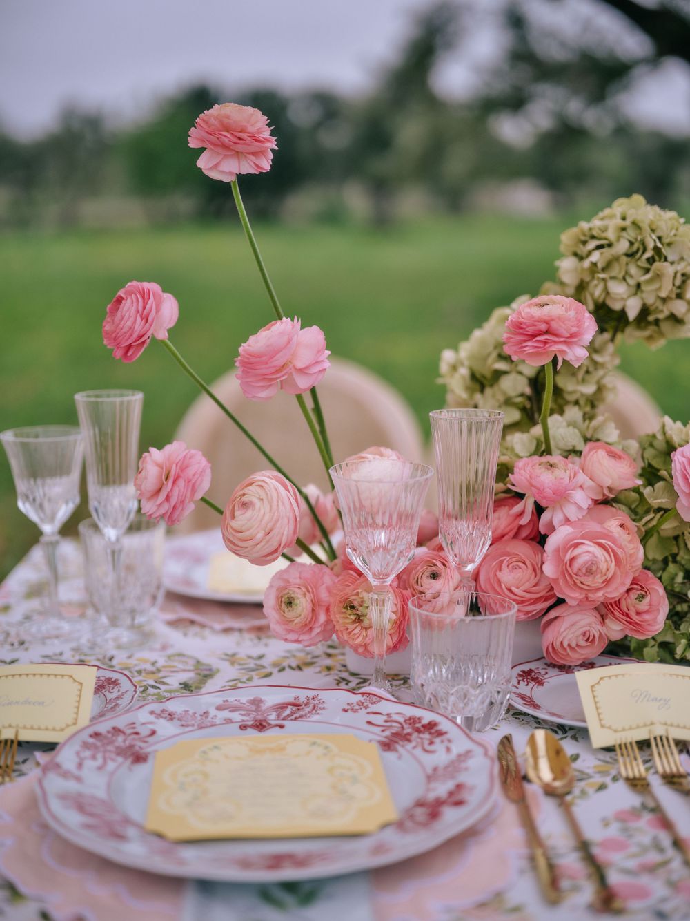 Elegant table setting with pink ranunculus flowers, floral plates, and crystal glasses on a delicate tablecloth.