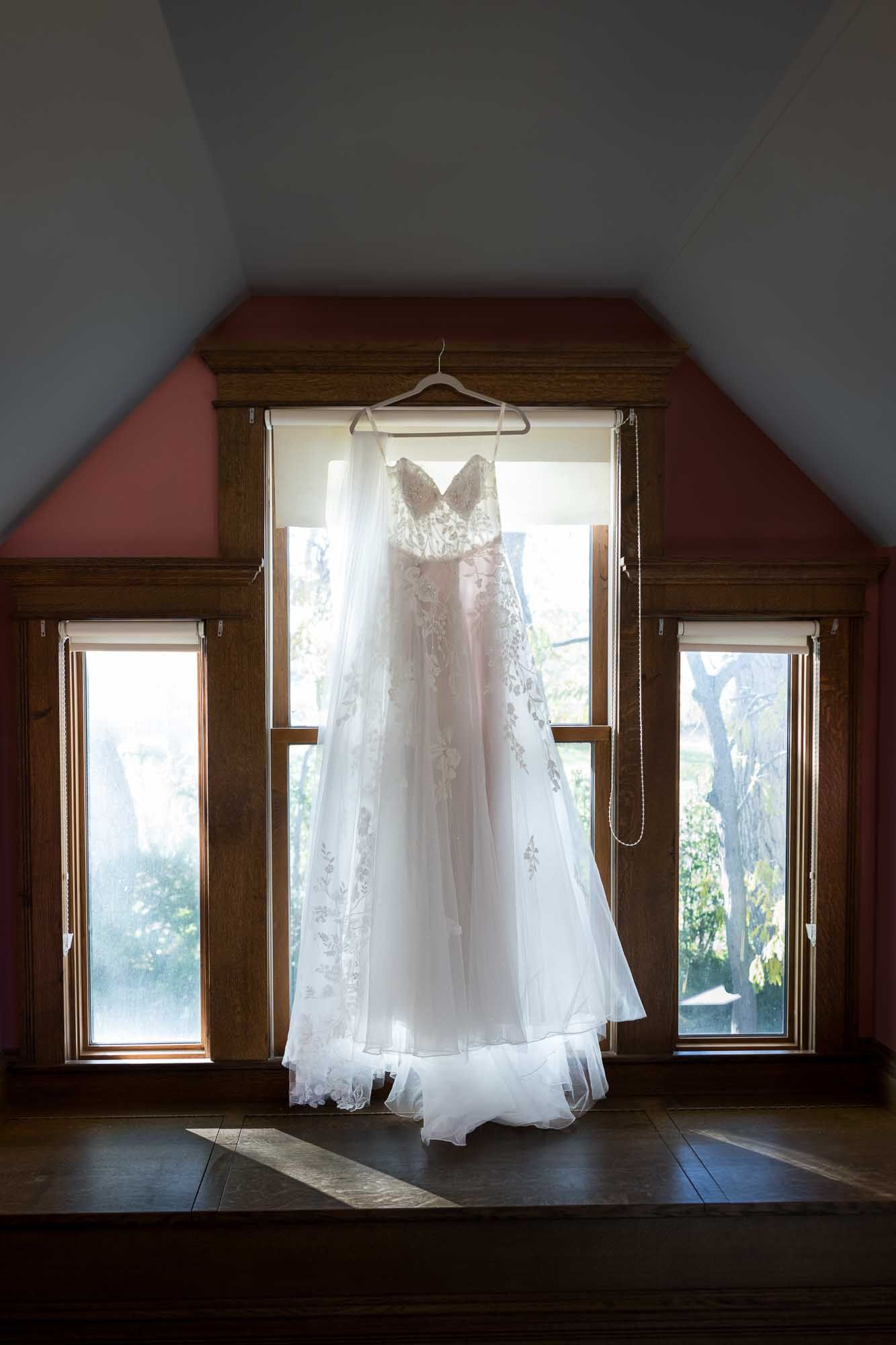 A delicate white lace wedding dress hangs in front of a window with wooden trim, backlit by natural daylight.