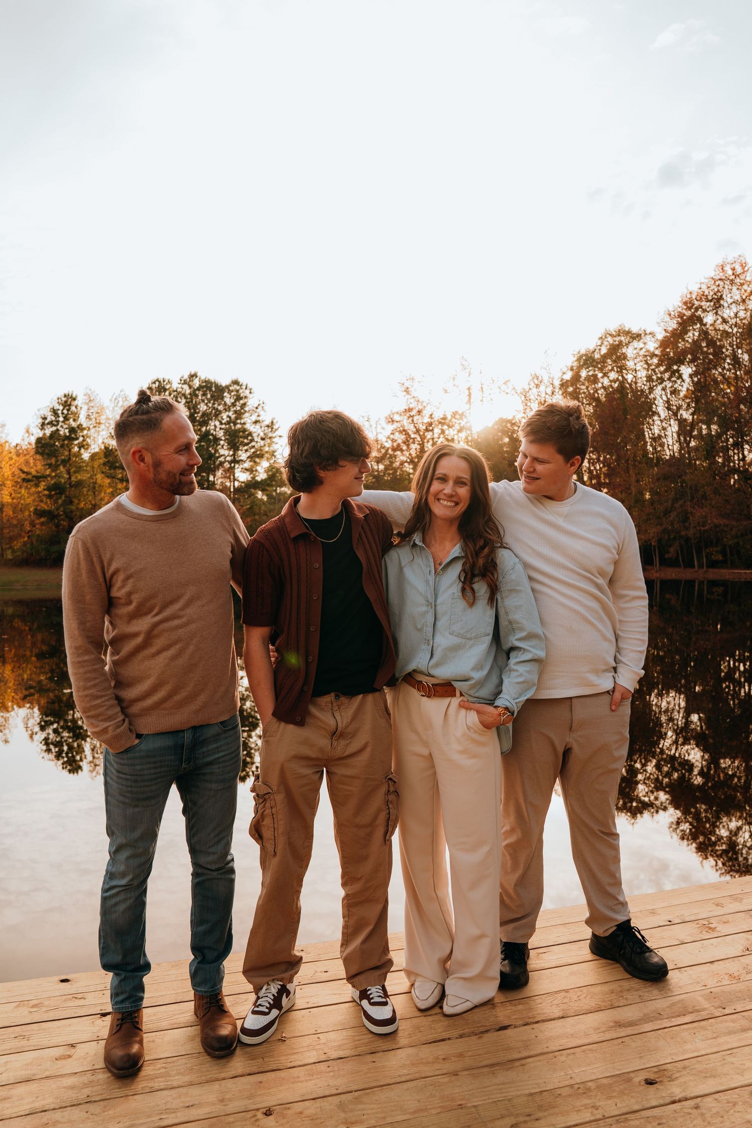 Family group stands together on wooden dock at sunset with lake and autumn trees in background.