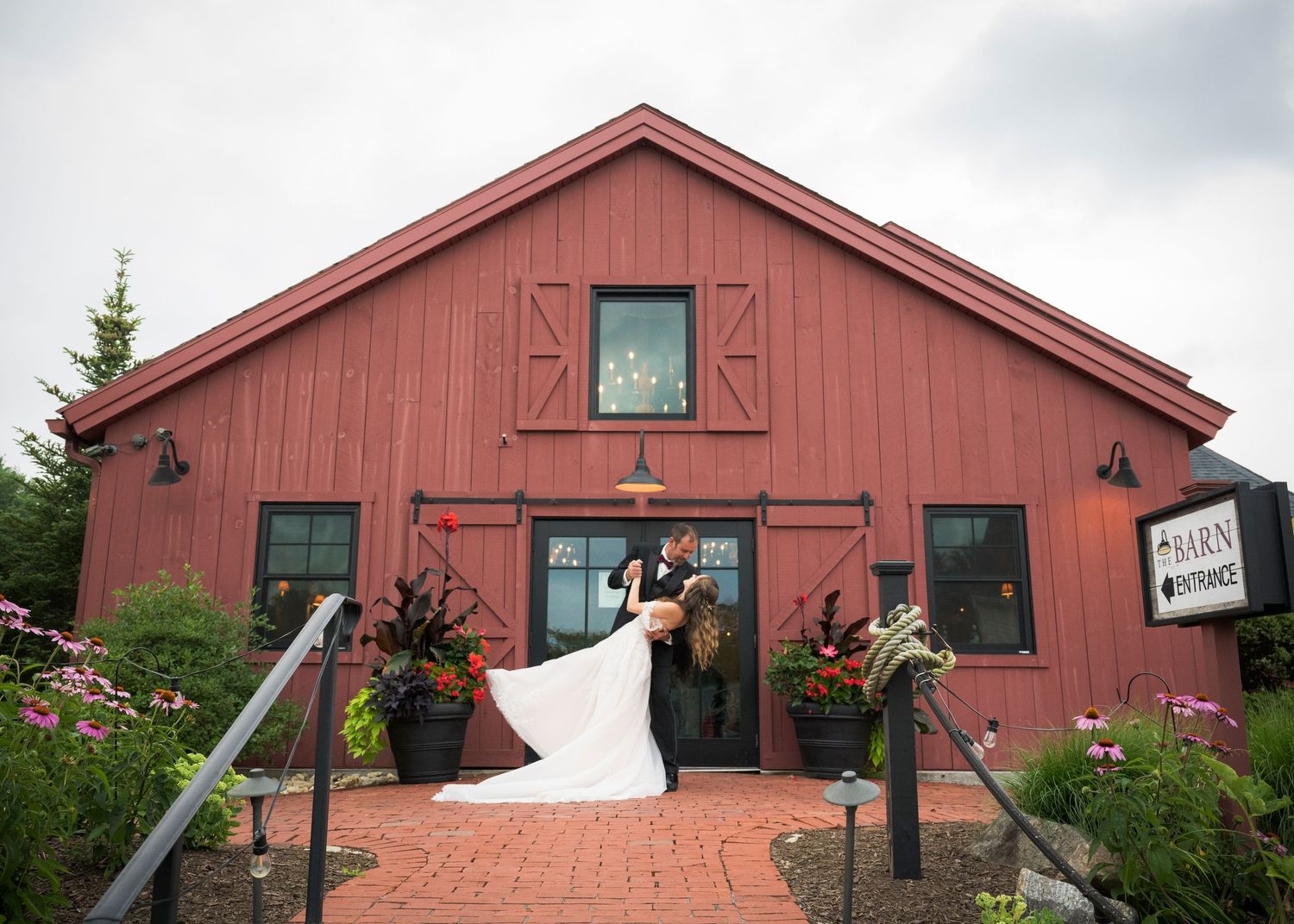 A panoramic view of a rustic red barn with black trim and windows against an overcast sky.