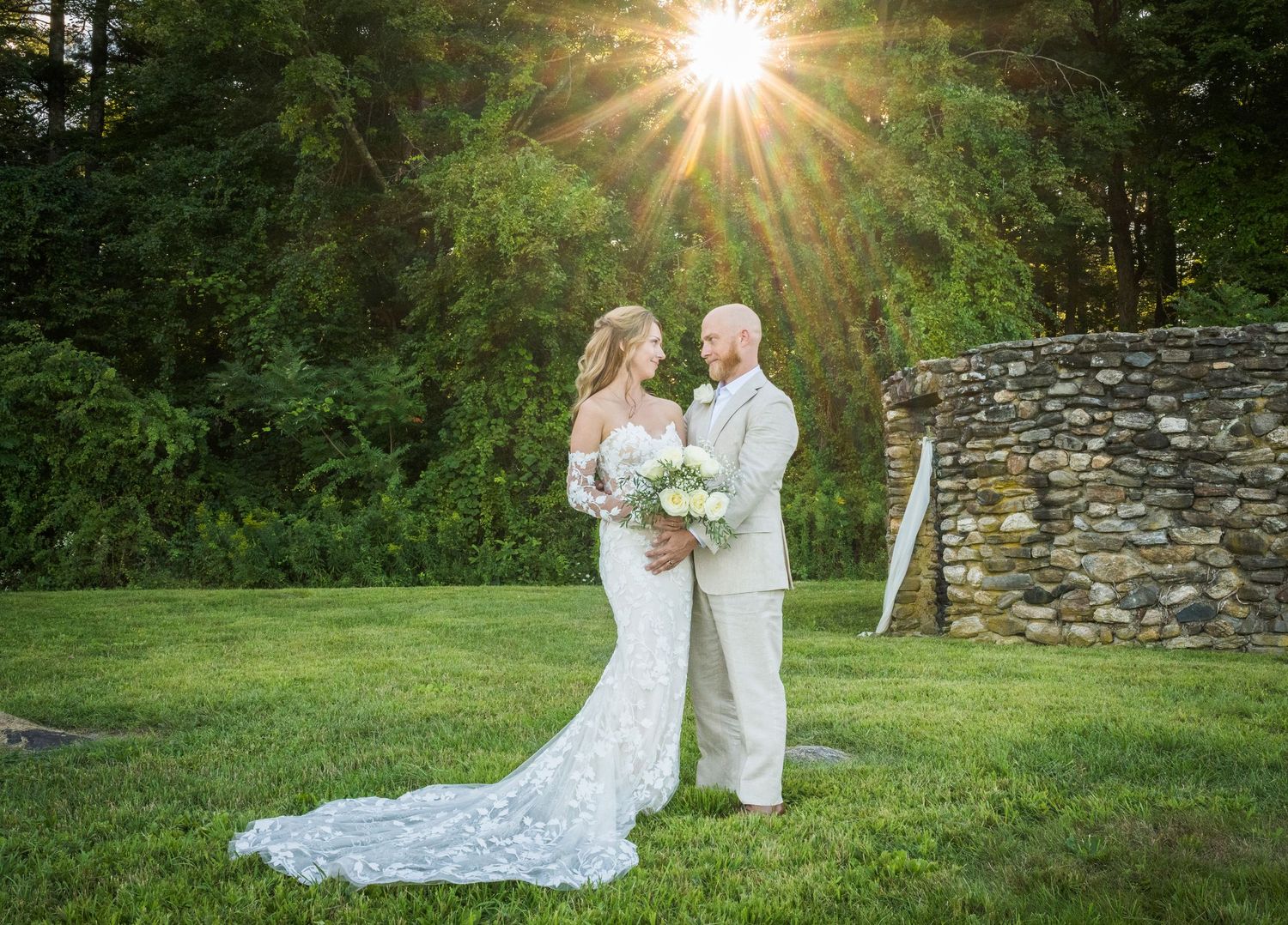 Sunburst through trees illuminates a romantic garden wedding scene with stone wall backdrop.