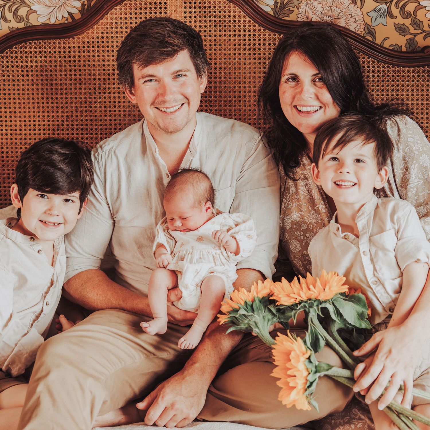 Family photo showing a couple with their 2 boys smiling at the camera, dad is holding their newborn baby girl.  They are sitting on a bed.