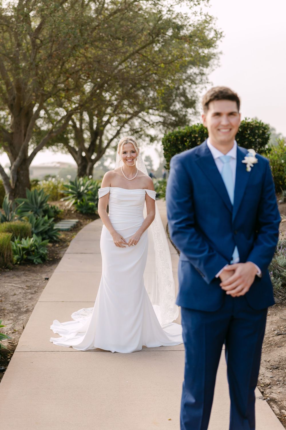 A couple in wedding attire stands together on a garden pathway with trees and landscaping in the background.