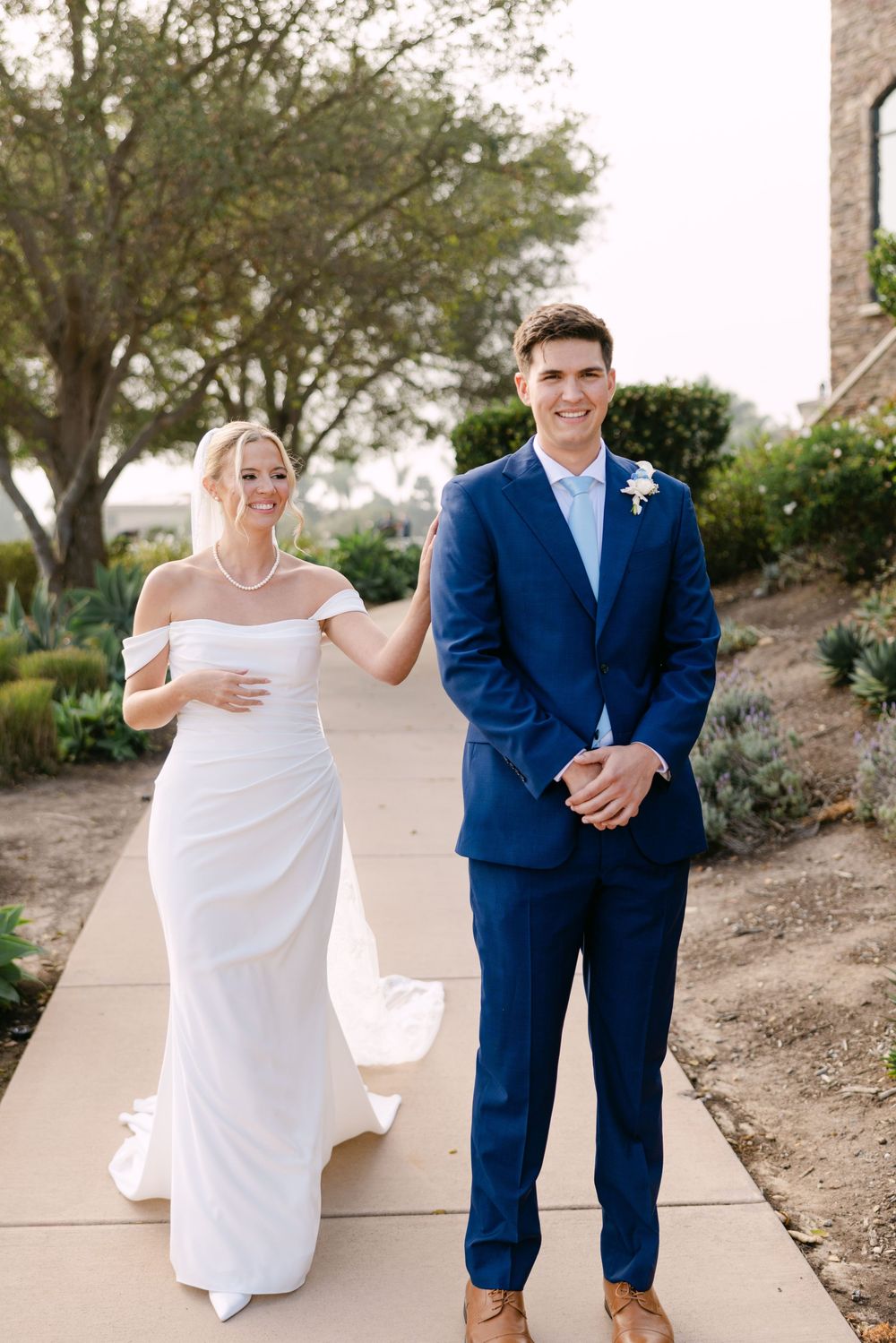A couple in wedding attire stand together on a path with a stone building and trees in the background.