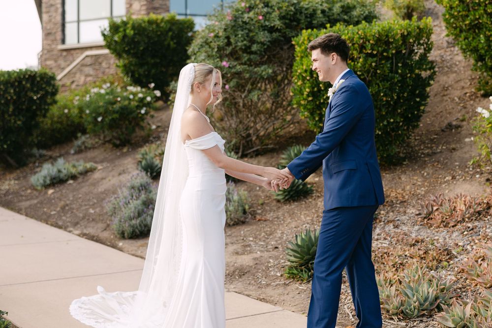 A couple in formal wedding attire share a romantic moment outdoors on a stone pathway surrounded by greenery.
