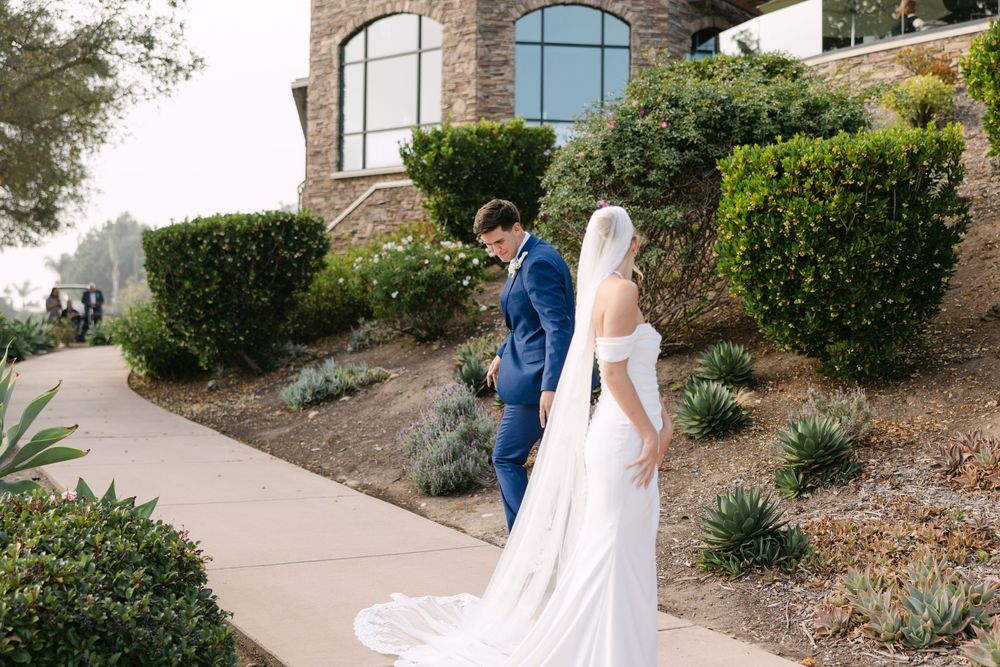 A couple in wedding attire walks along a winding stone path surrounded by lush landscaping and greenery.