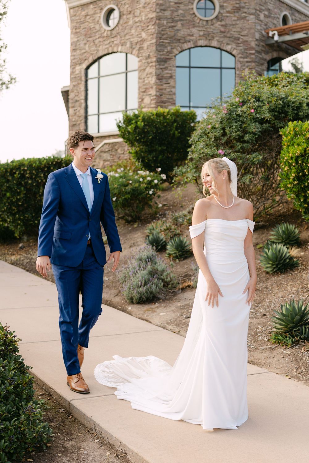 A couple in wedding attire walk along a path near a stone building with arched windows.