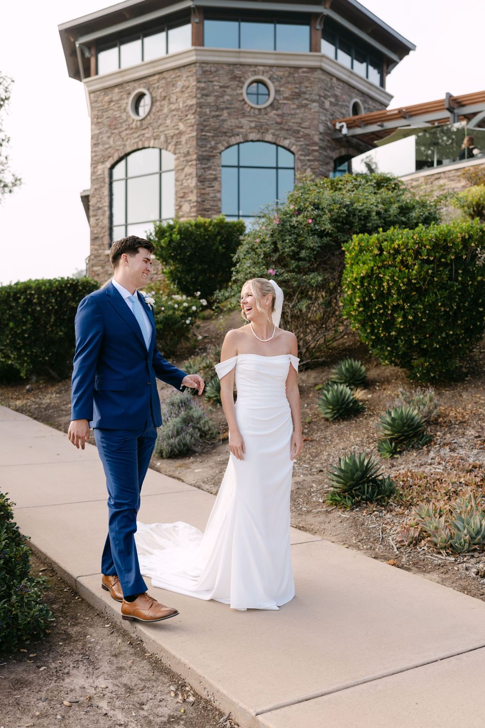 A couple in wedding attire share a joyful moment outside a stone building with round windows.