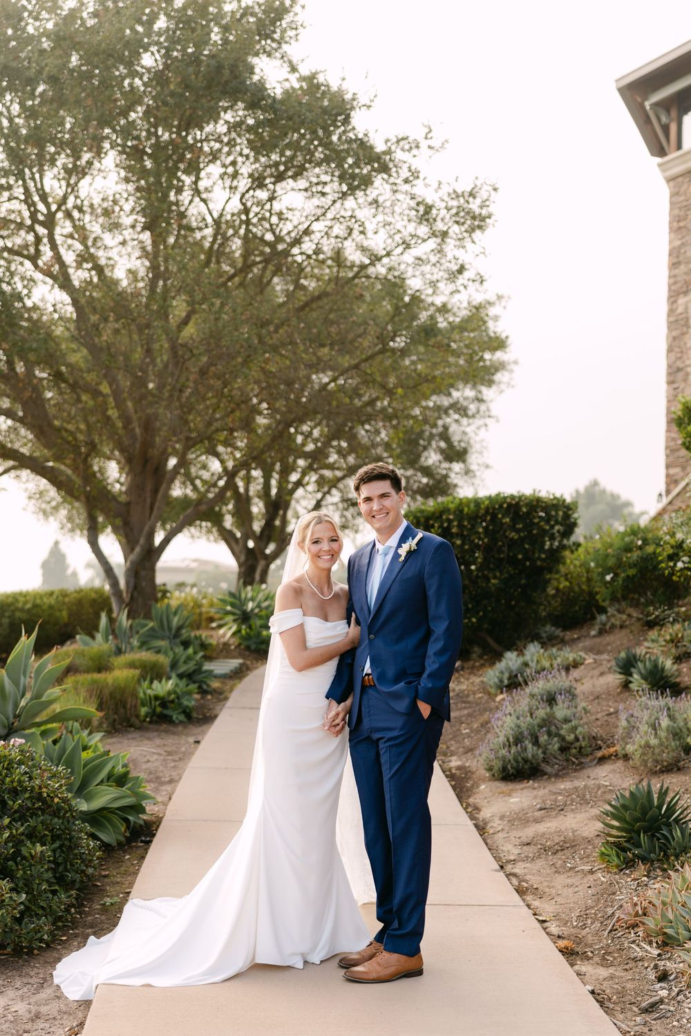 Wedding couple poses together on a landscaped pathway surrounded by desert plants and trees.