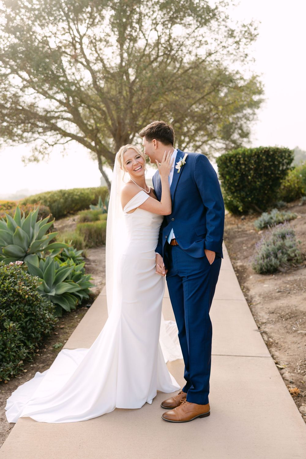 A couple in wedding attire share a romantic moment on a path lined with agave plants and desert landscaping.