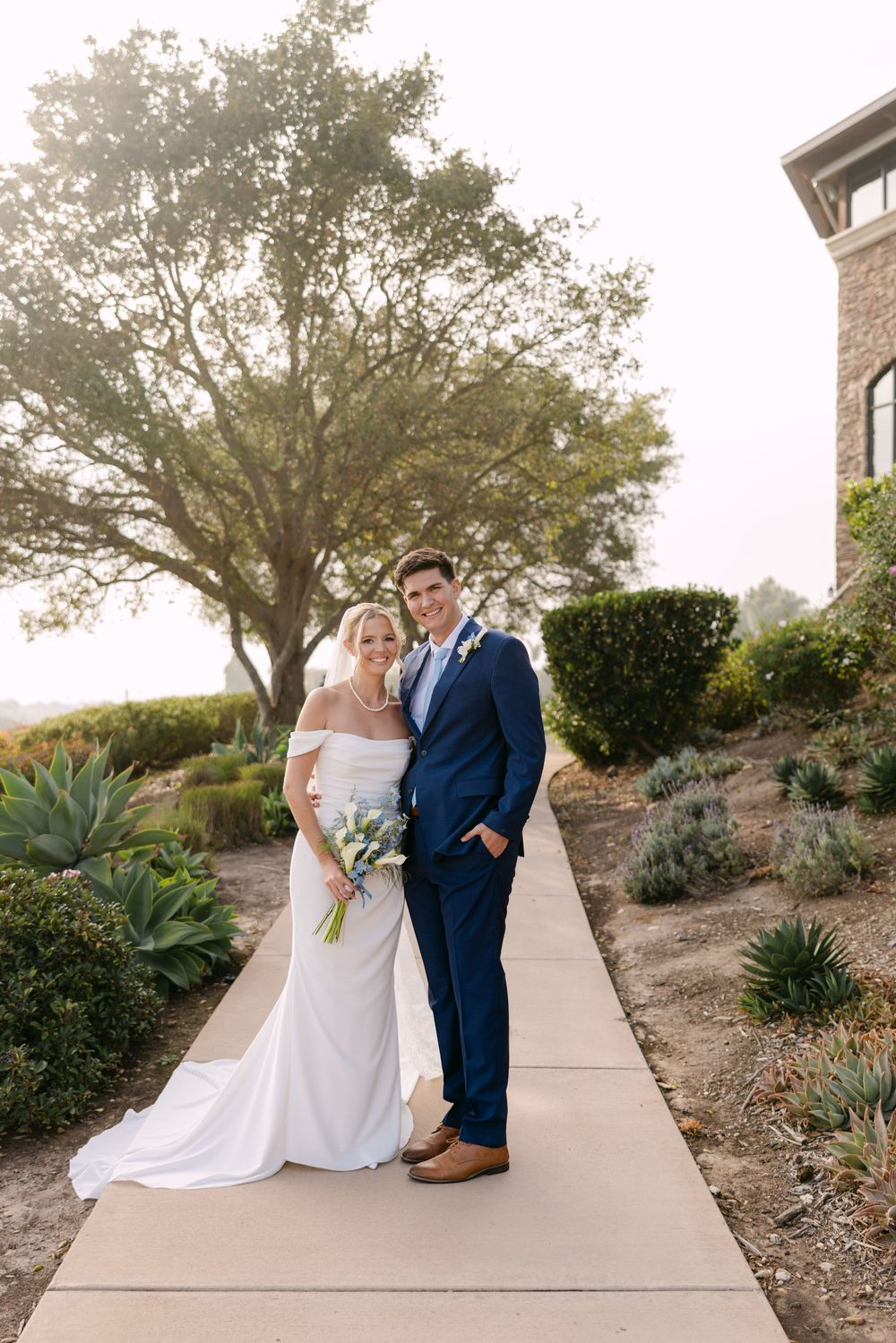 Wedding couple poses on a concrete walkway surrounded by desert landscaping and trees.