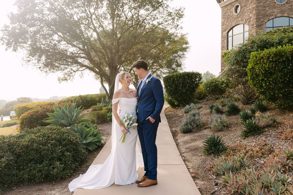 A couple in wedding attire pose together on a path with landscaped grounds and stone architecture.