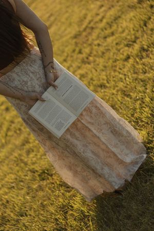 Woman in lace skirt holding open book while standing in sunlit golden grass field.