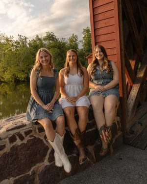 Three young women in denim outfits and cowboy boots sitting on stone steps near a red barn by a river.