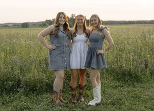 Three smiling women in summer dresses and cowboy boots posing together in a lush green field at sunset.