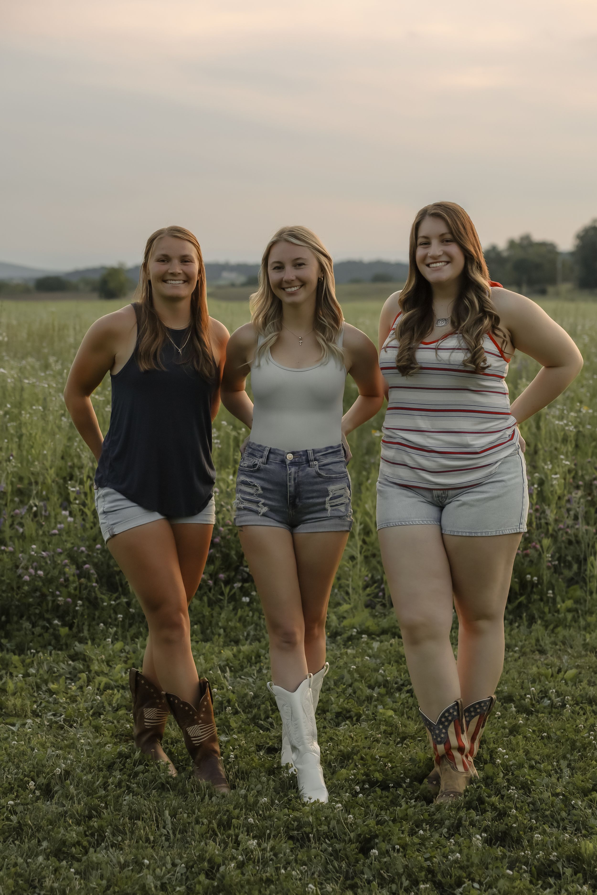 Three women in casual summer outfits and cowboy boots smiling together in a green field at dusk.