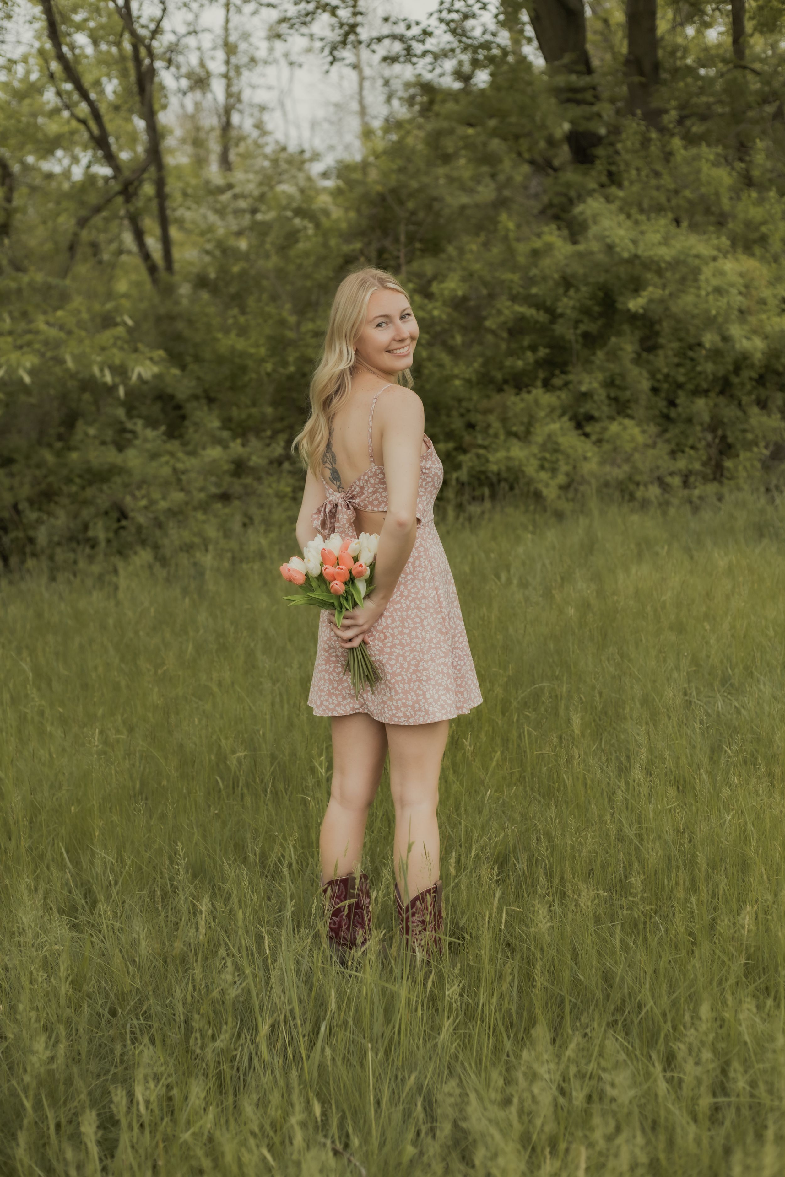 Smiling blonde woman in pink floral dress and cowboy boots holding flowers in lush green field.