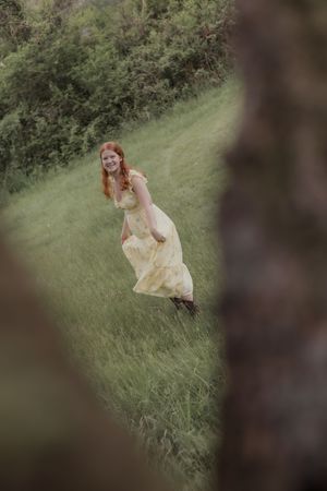 Redhead woman in yellow dress walking and smiling on green hillside path framed by tree.