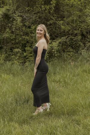 Smiling blonde woman in strapless black maxi dress standing in green field with wooded backdrop.