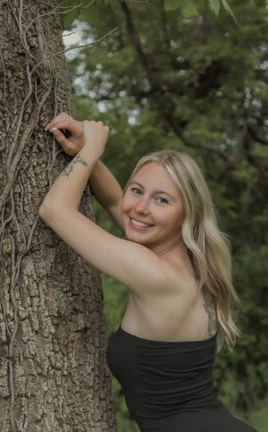 Smiling blonde woman leaning against tree trunk in black top surrounded by lush green forest.