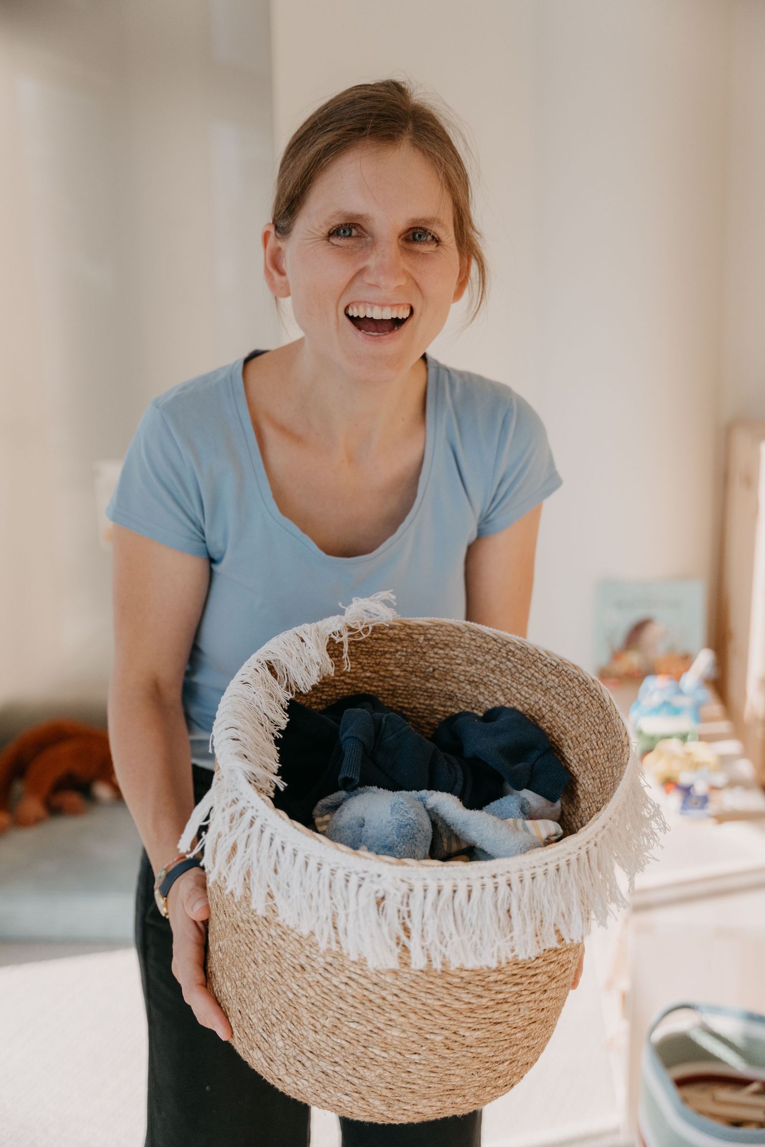 A person in a light blue shirt smiles while holding a round woven basket containing a small dark pet.