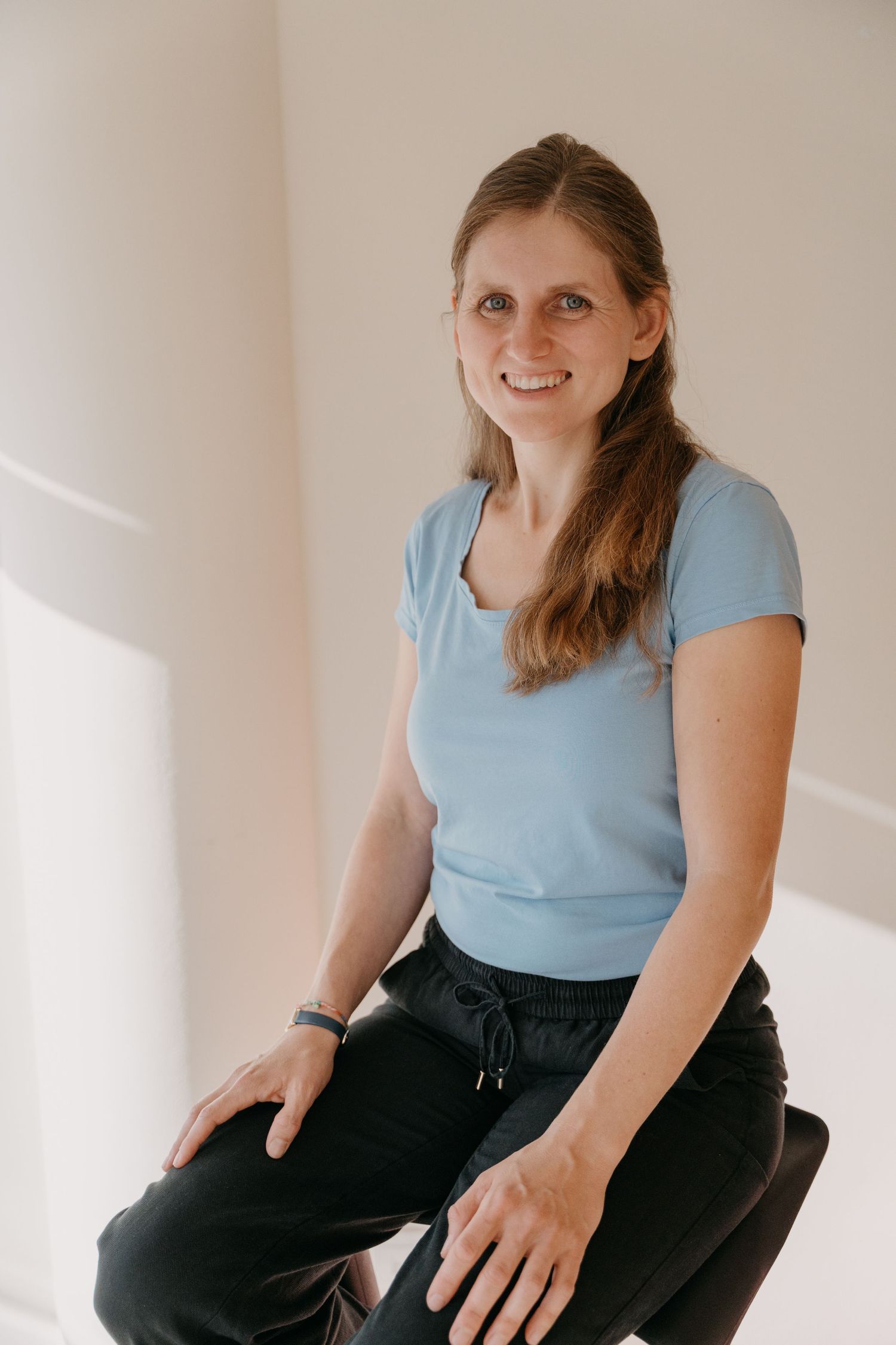 A woman in a light blue t-shirt and dark pants sits casually on a white surface while smiling warmly.